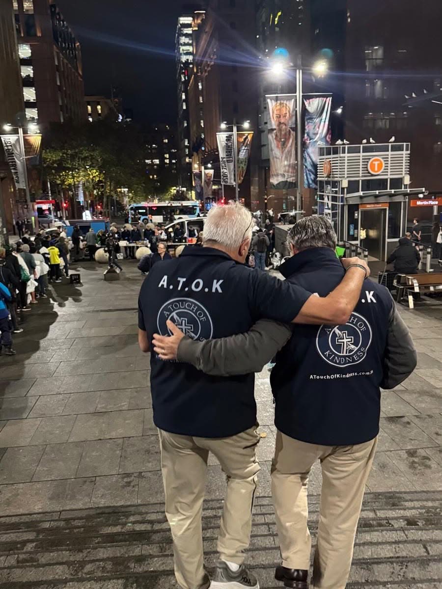 Two volunteers in A Touch of Kindness shirts with their arms around each other, walking through Martin Place at night